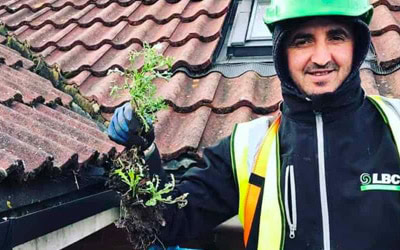 A man in a green helmet and high-visibility vest smiles as he holds up a weed he pulled from a building’s rain gutter. The background shows a tiled rooftop and part of a structure. The man appears to be involved in gutter cleaning or maintenance work.