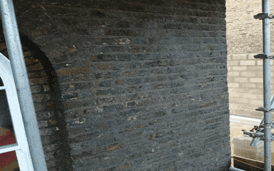 Commercial, A brick wall with a curved archway undergoes renovation. The archway is filled with matching bricks, making it appear as a solid wall. Scaffolding is present on the right, and a partially visible white structure is on the left. The scene suggests an ongoing construction or restoration project.