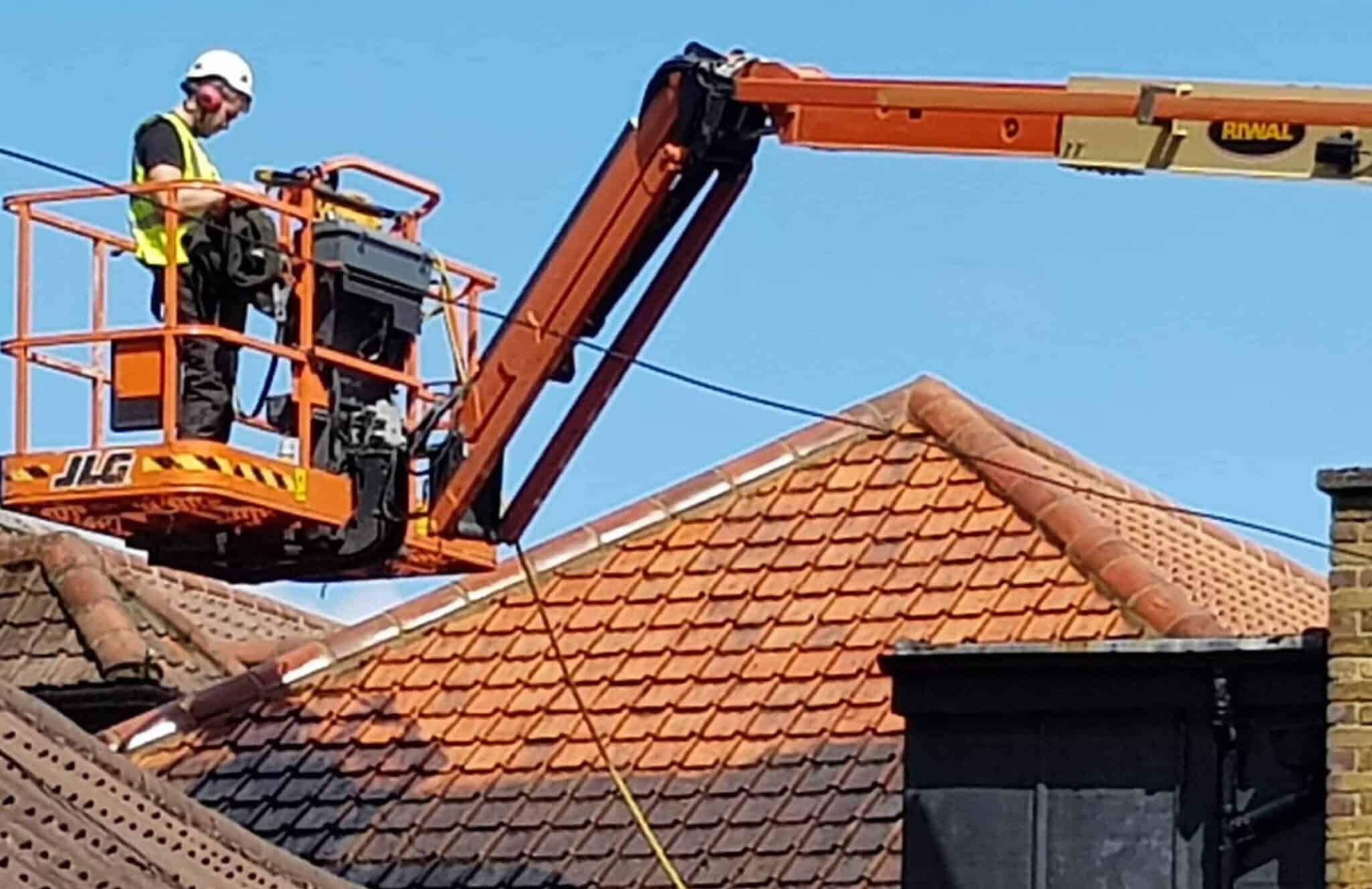 A worker in a neon yellow safety vest and helmet operates an orange JLG cherry picker lift to perform maintenance on brown and red roof tiles of a building. The clear blue sky serves as the backdrop, and a brick chimney is partially visible on the right side of the image.
