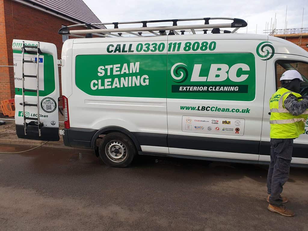 A white van with green branding for LBC Exterior Cleaning, featuring the text "STEAM CLEANING" and "CALL 0330 111 8080." The van has ladders on the roof and various logos on its side. A person in a safety vest and helmet stands beside the van on a paved road.