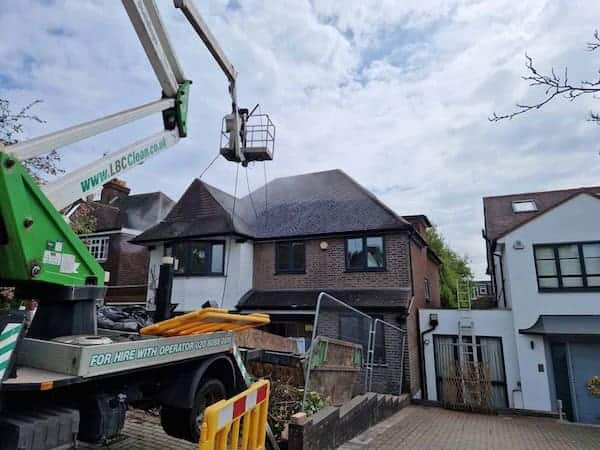 A worker in a hydraulic lift power washes the roof of a two-story brick house. The lift has "LBClean.co.uk" printed on its arm. The house is flanked by similar houses, and construction materials are visible on the driveway, which is partially blocked by a safety fence. The sky is partly cloudy.