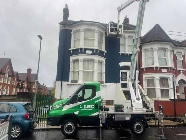 A green and white LBC utility vehicle with an elevated work platform is parked in front of a row of Victorian-style houses on a rainy day. The platform extends to the roof of a navy blue and white house where a worker can be seen performing maintenance. A blue car is parked nearby.