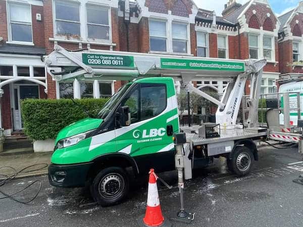 A green and white LBC exterior cleaning truck with a mounted telescopic lift is parked on a residential street in front of brick row houses. There is a traffic cone and some cleaning equipment on the road next to the truck. LBC's contact details are visible on the truck's side.