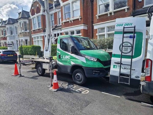 Green and white GB LBC truck parked in a residential street, performing utility work. Orange traffic cones placed around the vehicle. Brick houses with plaster decorations and white trim in the background. Truck features the company logo and contact number on the rear and side.