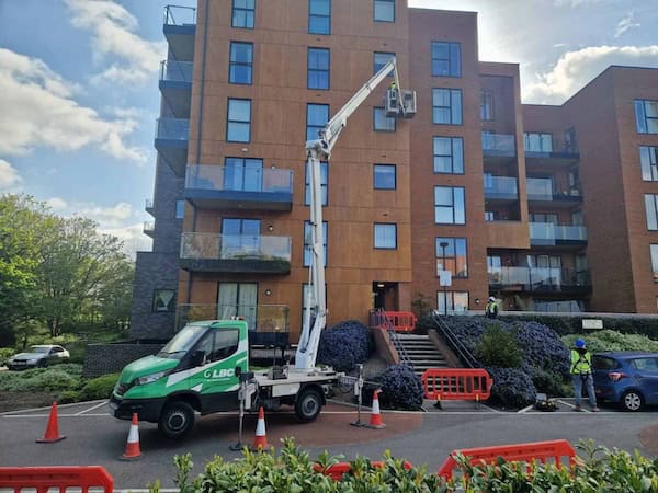 A green LBC utility truck with an elevated platform is parked in front of a multi-story apartment building. Workers in bright vests are operating the platform near a balcony. Orange safety barriers and cones surround the area. The sky is partly cloudy, and the building is surrounded by greenery.