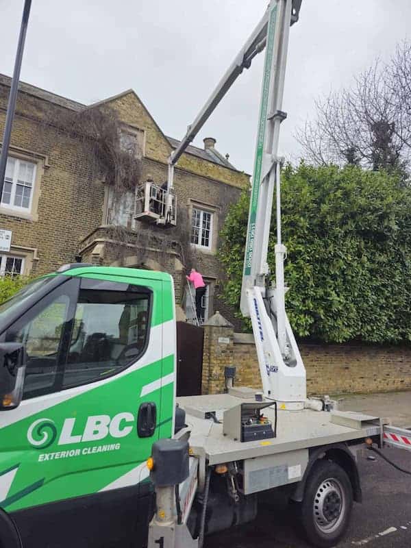 A worker in a pink jacket stands in a cherrypicker lift raised to the second-story level of a brick building, cleaning the exterior. The LBC-branded cherrypicker truck is parked on the street below. The building has white-trimmed windows and is partially obscured by an overgrown bush.