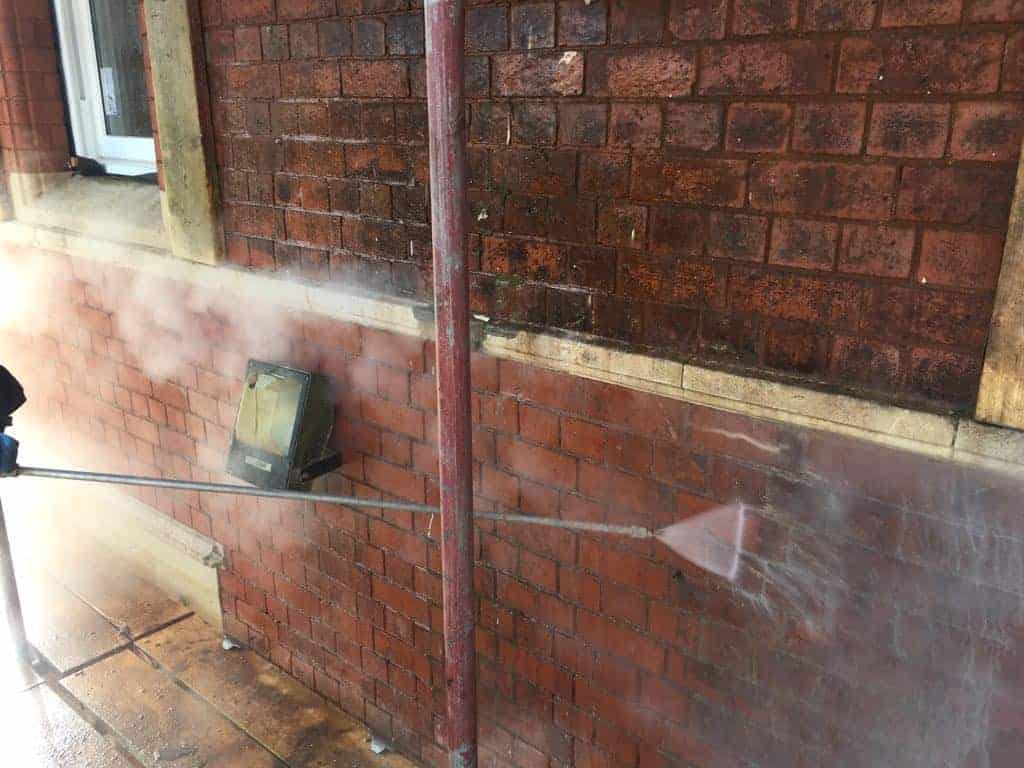 A person power washes a brick wall, removing dirt and grime. The wall, brownish-red in color, shows a clear contrast between the cleaned section and the still-dirty part. Steam rises from the area being cleaned. A wooden window frame and a concrete floor are partially visible.