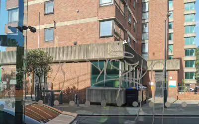 A brick building with a flat roof is tagged with graffiti in white paint. The exterior has several windows, a few of which are partially open. There is a small tree and a dumpster near the building's entrance. The scene appears to be on a sunny day, with shadows cast on the ground, suggesting potential need for exterior cleaning services.