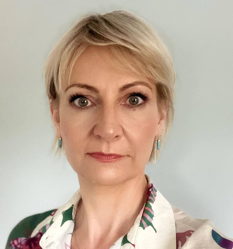 A person with short blonde hair from the LBC team looks directly at the camera with a neutral expression. They are wearing a floral-patterned shirt and small turquoise earrings, set against a plain, light-colored backdrop.