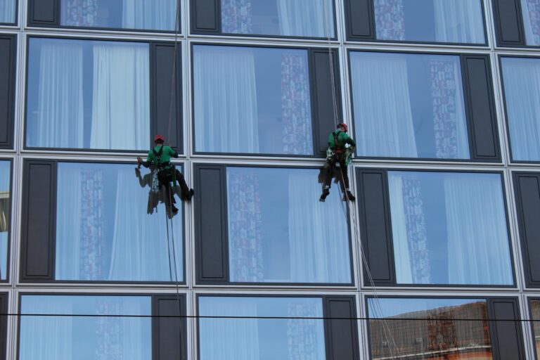 Two window washers in green uniforms and red helmets perform abseil window cleaning, suspended by ropes as they clean the glass of a modern multi-story building with large square windows and some curtains visible inside.