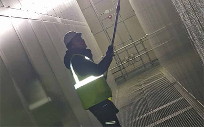 A person in a hard hat, high-visibility vest, and dark clothes uses an extended tool to clean or inspect a tall wall inside a commercial industrial facility with metal grating floors and large vertical panels under industrial lighting.