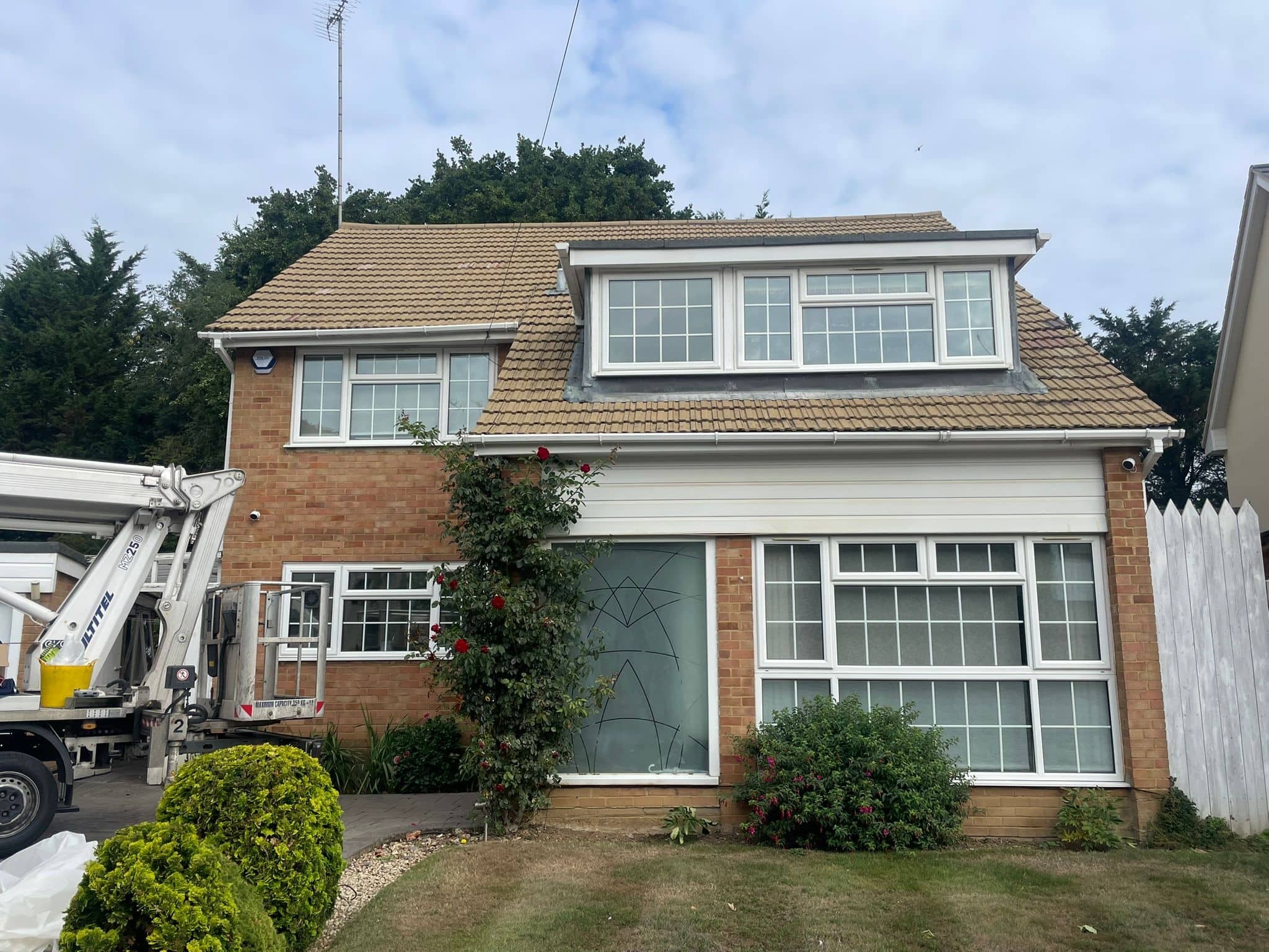 A two-story brick house with large front windows, a pitched tan roof, and a glass door partially covered by a leafy vine with red flowers. A white utility truck—possibly for Roof Spraying & Recoating Services—is parked to the left. Trimmed bushes line the patchy lawn.
