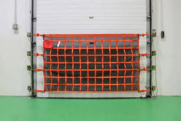 A bright orange safety barrier net is installed in front of a white industrial garage door, secured with metal brackets. The floor is painted green, the walls are plain white with some visible electrical outlets, and a bird figure decorates one corner.