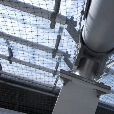 Close-up of a metal structural beam connected to a cylindrical support, viewed from below. Bird-safe protective mesh netting is stretched above, with sunlight filtering through transparent roof panels in the background.