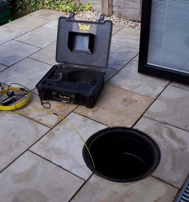 A black Specialist Rat Investigation & Proofing inspection camera case with a monitor and keyboard sits on wet paving stones next to an open manhole. A yellow cable extends from the case, while glass doors and a garden with rocks and plants are nearby.